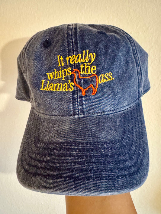 A dark blue acid-wash denim baseball cap with yellow and orange embroidered text reading 'It really whips the Llama's ass.' and an orange llama outline, shown against a plain beige wall.